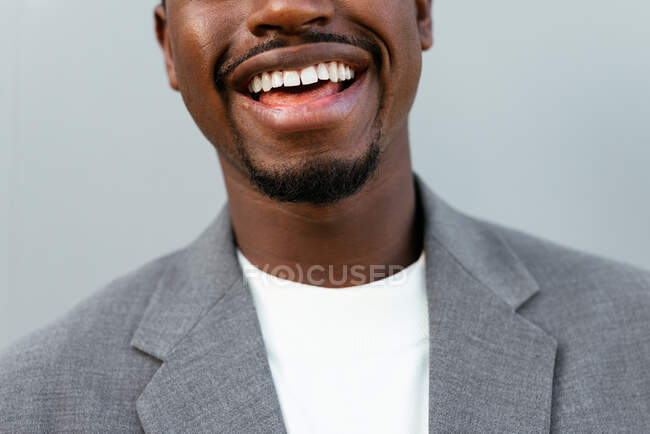 Crop entrepreneur afro-américain masculin en costume formel souriant largement tout en se tenant debout sur fond gris et en regardant la caméra — Photo de stock