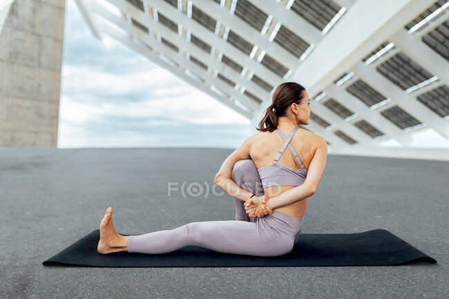 Ganzkörper-Rückenansicht einer nicht wiederzuerkennenden sportlichen Frau in Aktivkleidung, die auf einer Matte sitzt, während sie Yoga-Asana mit Twist in der Nähe von Solarzellen auf der Straße praktiziert — Stockfoto