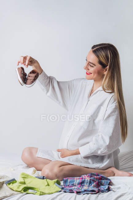 Positive junge Frau im weißen Hemd sitzt auf dem Bett und bereitet Babykleidung der Geburt vor, während sie auf dem Bett sitzt — Stockfoto