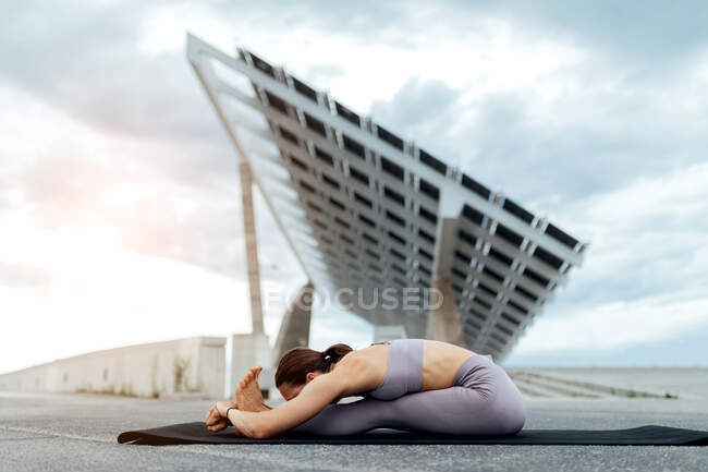 Cuerpo completo de mujer deportiva en ropa deportiva que practica la postura sentada hacia adelante mientras se entrena en la calle cerca del panel solar contra el cielo nublado - foto de stock