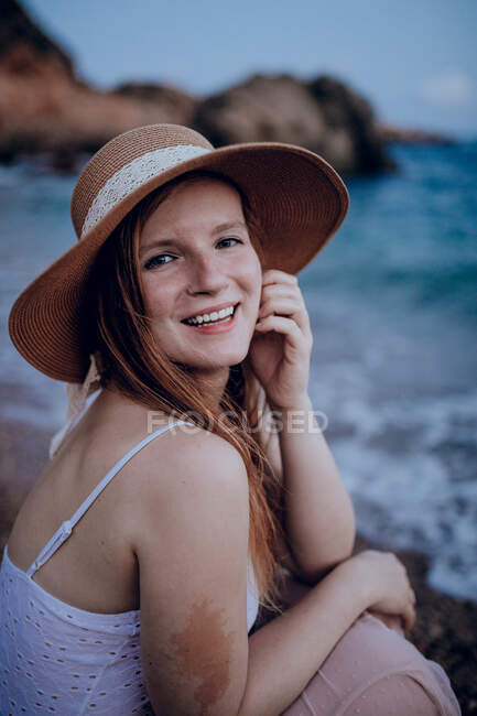 Обаятельная женщина в шляпе, сидящая на пляже у моря и трогающая лицо, улыбаясь и глядя в камеру летом — стоковое фото