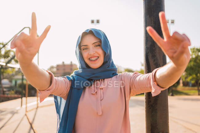 Delighted Arab female in traditional headscarf showing two fingers gesture and looking at camera while standing in city on sunny day — Stock Photo