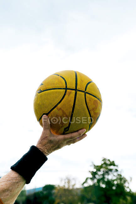 Cosecha anónima persona madura con pelota de baloncesto amarillo en la mano de pie en el campo de deportes públicos mientras juega juego en la calle - foto de stock