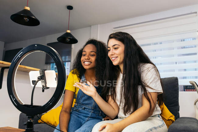 Mujer positiva con el pelo largo y oscuro en ropa casual sentada y mostrando en el alegre blogger afroamericano mientras graba vlog en un teléfono inteligente moderno colocado en el trípode con lámpara de anillo led en casa - foto de stock