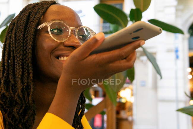 Mulher afro-americana positiva com cabelo preto em óculos de sol gravando mensagem de voz no smartphone moderno enquanto estava sentada no café com plantas — Fotografia de Stock