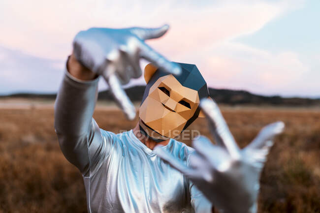 Unrecognizable person with geometric monkey mask demonstrating rock gesture on blurred background of rural land — Stock Photo