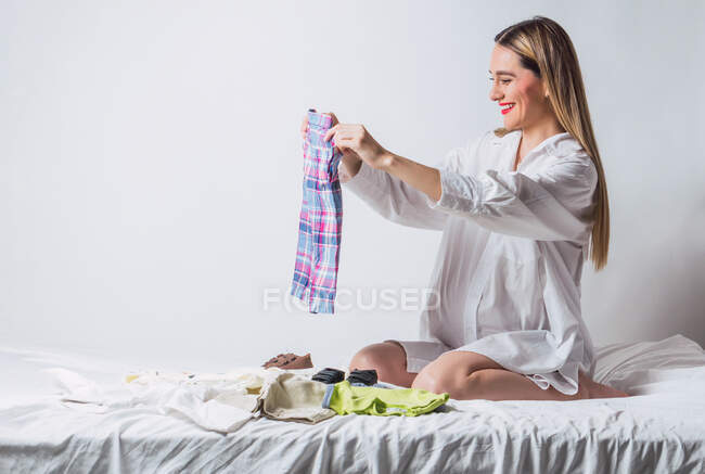 Cultivo embarazada hembra con labios rojos anticipando bebé plegable ropa de los niños mientras está sentado en la cama en el dormitorio - foto de stock