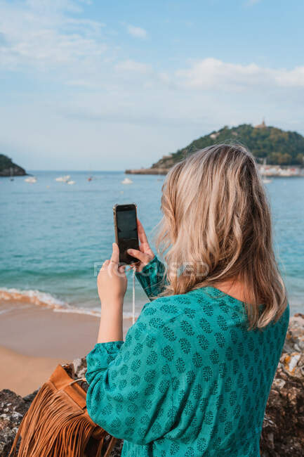 Vista lateral da elegante fêmea irreconhecível com cabelo loiro tirando foto do oceano azul contra a paisagem urbana de Donostia no smartphone durante o dia — Fotografia de Stock