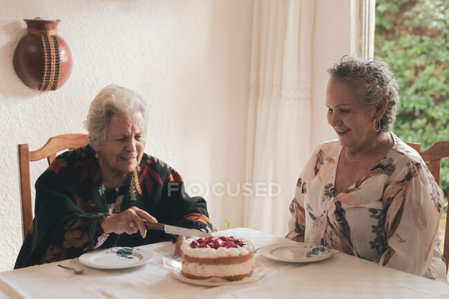 Mujer mayor sonriente en chal caliente con amiga sentada a la mesa y cortando deliciosa torta con bayas para el té - foto de stock