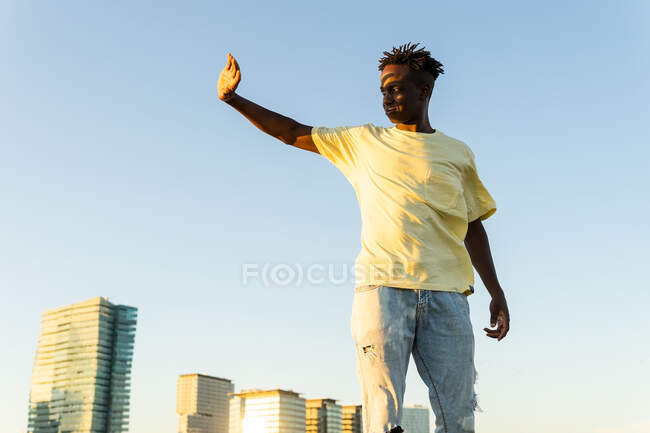 Low angle of young African American male in trendy outfit standing with raised arm on city street in summer evening — Stock Photo