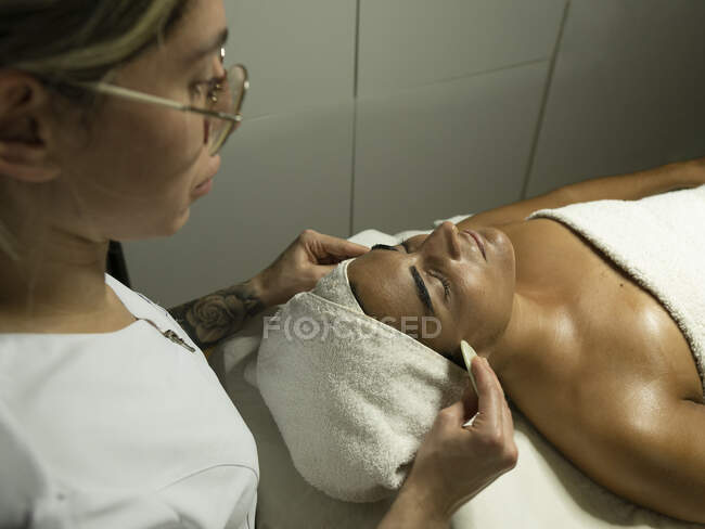 From above of female masseur using massagers while massaging face of client in towel on couch in bright spa center — Stock Photo