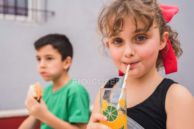 Bambini allegri che ridono e mangiano panini freschi seduti a tavola e bevono succo di frutta nella stanza luminosa di casa — Foto stock