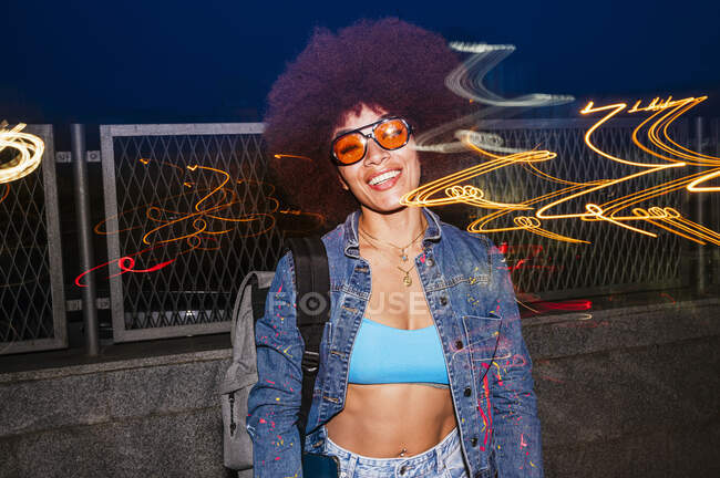 Cheerful female with sunglasses looking at camera while standing near fence with light effect in evening time — Stock Photo