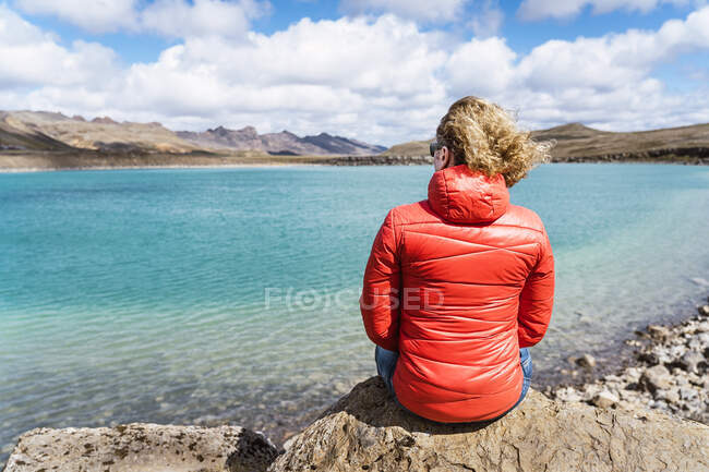 Indietro vista della calma viaggiatore irriconoscibile seduto sul bordo della scogliera sopra l'oceano blu contro le montagne in Islanda nella giornata di sole — Foto stock