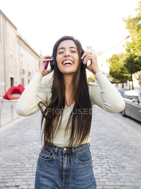 Cheerful young woman with long brown hair in casual clothes standing while listening to music in headphones on city street in daylight — Stock Photo