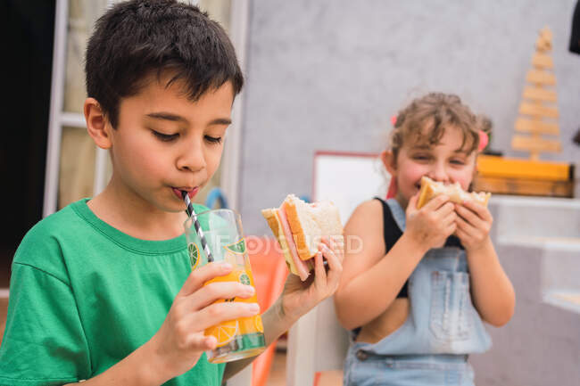Fröhliche Kinder lachen und essen frische Sandwiches, während sie zu Hause am Tisch sitzen und Saft trinken — Stockfoto