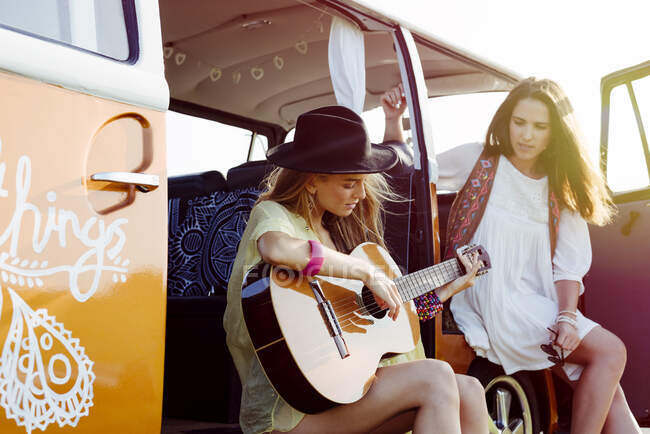 Дві красиві дівчата, одягнені в літній одяг, розважаються поза фургоном і грають на гітарі — стокове фото