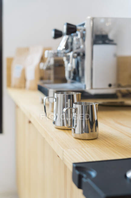 Foco suave de jarros profissionais inoxidáveis para derramar leite colocado no balcão de madeira na moderna casa de café com máquinas de cafeteira — Fotografia de Stock