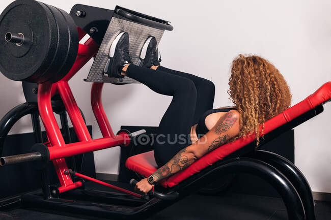 Seitenansicht einer nicht wiedererkennbaren Sportlerin mit lockigem Haar, die im Fitnessstudio Übungen auf einem Beinpressgerät macht — Stockfoto