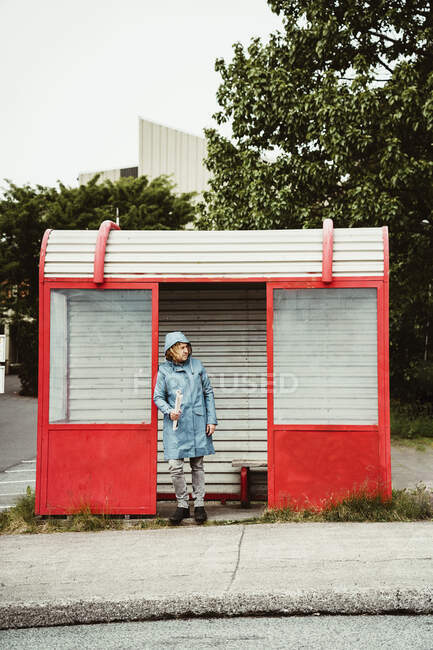 Полное тело задумчивого человека в дождевике, стоящего и опирающегося на стену автобусной остановки возле проезжей части в городе при дневном свете — стоковое фото