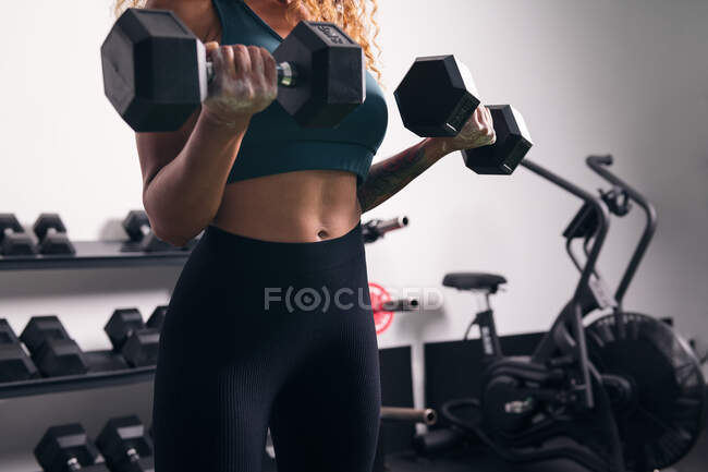Crop strong sportswoman with curly hair doing exercise on biceps with dumbbell during workout in gym — Stock Photo