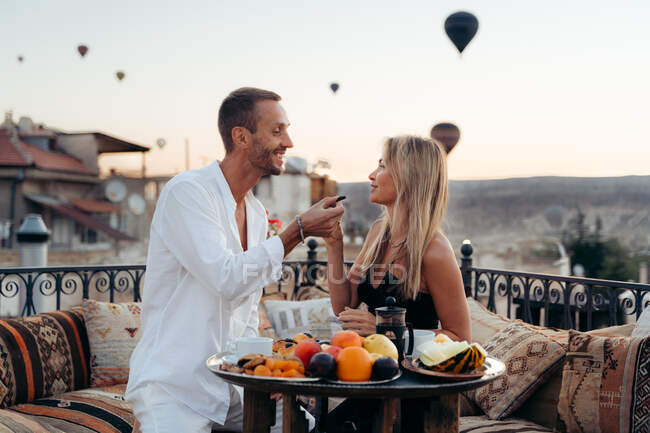 Paar hält Händchen und schaut einander beim romantischen Abendessen an und füttert sich gegenseitig mit Früchten auf der Terrasse mit Heißluftballons im Hintergrund in Kappadokien — Stockfoto