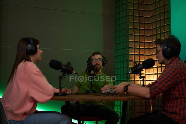 Colegas y colegas jóvenes concentrados en ropa casual y auriculares sentados en la mesa con micrófonos y comunicándose mientras graban podcast en estudio moderno - foto de stock