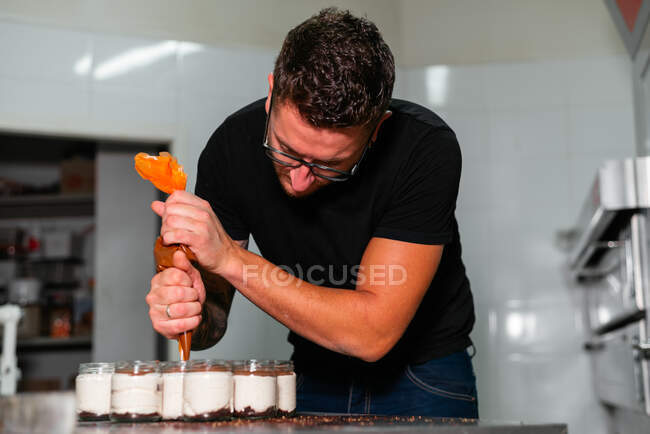 Attentive young male baker in casual clothes and eyeglasses adding caramel topping from pastry bag into jars with delicious layer dessert in kitchen — Stock Photo