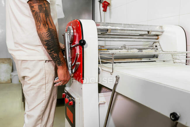 Вид сбоку мужской шеф-повар в повседневной одежде, стоящий и работающий на тестопрокатной машине при приготовлении теста на профессиональной кухне — стоковое фото