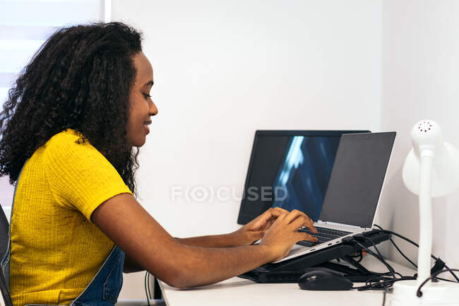 Vista lateral do conteúdo Mulher afro-americana sentada à mesa e digitando no laptop na sala de luz — Fotografia de Stock