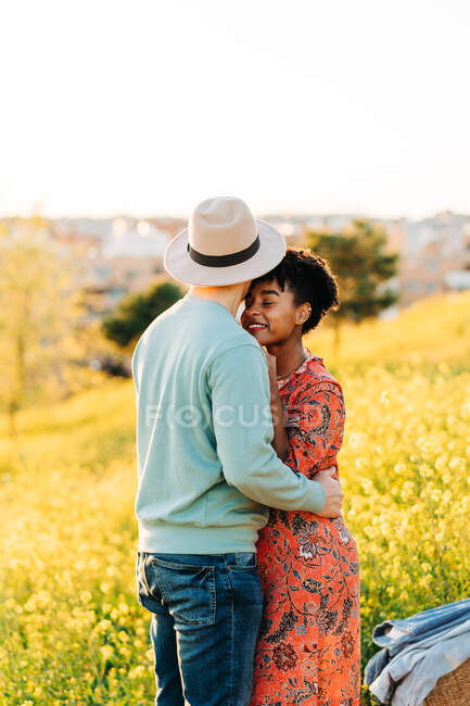 Vista lateral de la joven mujer afroamericana feliz con el pelo corto y oscuro sonriendo mientras abraza novio anónimo con los ojos cerrados de pie en el prado floreciente en el día soleado - foto de stock
