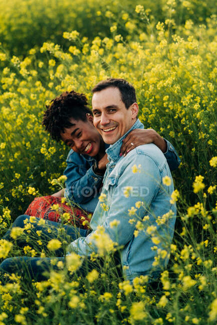Amar jovem casal multirracial em roupas casuais olhando para a câmera e sentado no prado florescendo exuberante durante a data romântica no dia ensolarado — Fotografia de Stock