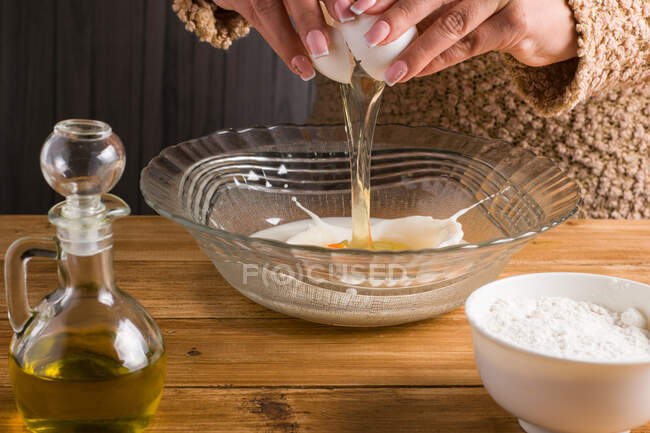 Cultivar hembra anónima rompiendo huevo crudo en leche en la mesa con aceite y harina mientras se cocinan crepes en la cocina ligera - foto de stock