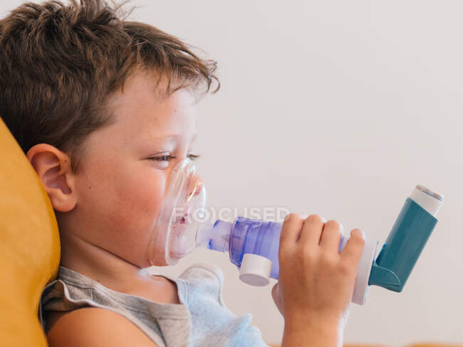 Vista lateral del niño enfermo contenido usando respirador y oxígeno respiratorio del inhalador mientras está sentado en el sofá en casa - foto de stock