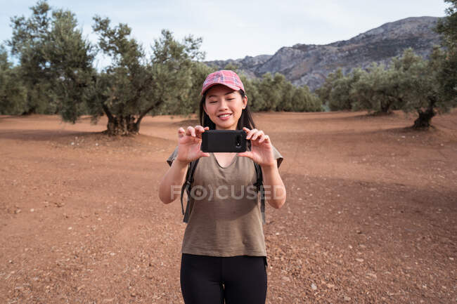 Sonriente hembra asiática en gorra tomando fotos en un campamento de olivo verde con un teléfono inteligente moderno mientras está de pie en el bosque - foto de stock