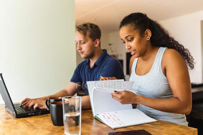 Coppia multietnica che lavora su progetto a distanza da casa insieme utilizzando laptop annotare note in copybook seduto al tavolo della cucina — Foto stock
