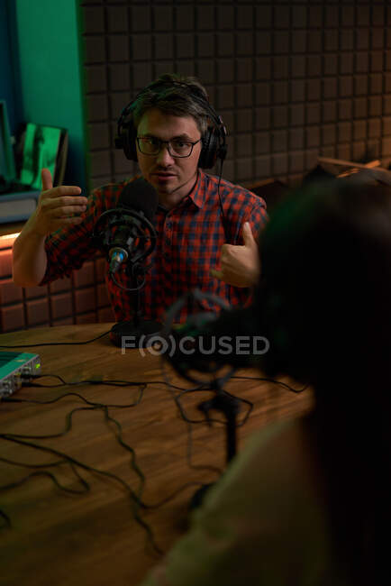 Jeune animateur radio masculin en chemise à carreaux et écouteurs assis à table avec microphone et communiquant avec un collègue anonyme lors de l'enregistrement de podcast en studio — Photo de stock