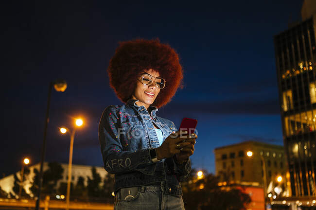 Позитивна жінка з африканською зачіскою та сучасним текстом на мобільному телефоні, стоячи на вулиці з будинками та ліхтарями у вечірній час. — стокове фото