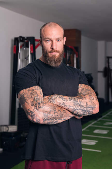 Fuerte deportista adulto con barba y tatuajes en brazos cruzados mirando a la cámara en el gimnasio - foto de stock