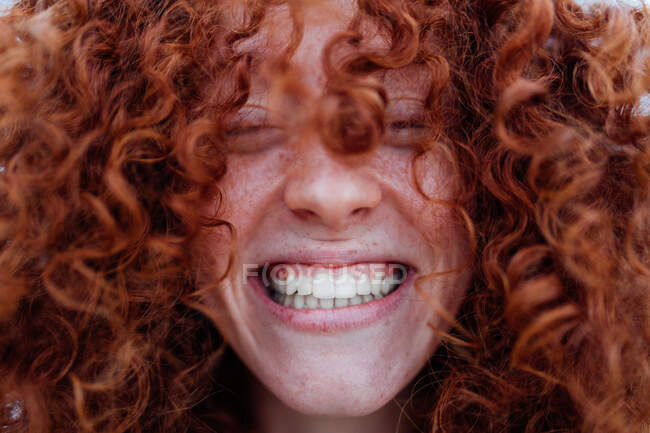 Delighted young female covering face with curly red hair while laughing happily with closed eyes — Stock Photo