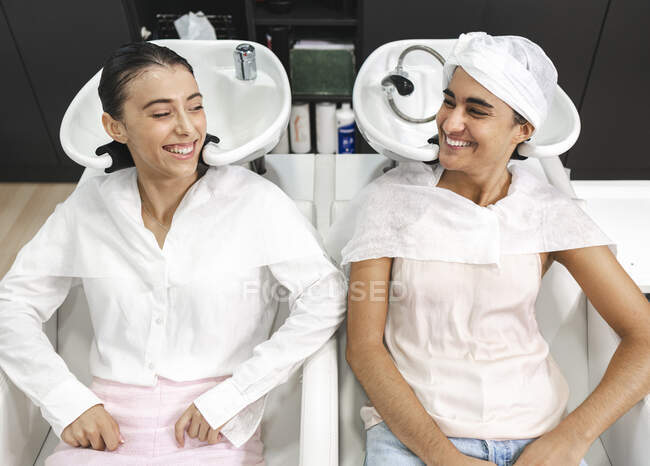 Desde arriba de las hembras sonriendo y mirándose entre sí mientras se sientan en sillas en el salón de peluquería después de lavar el cabello - foto de stock