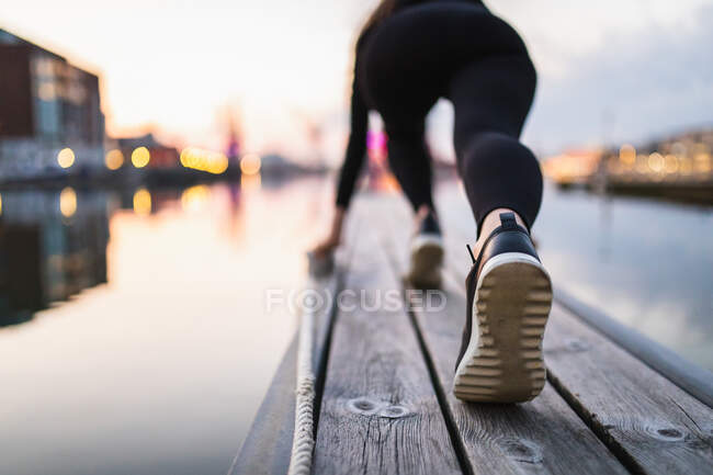 Вид сзади полное тело спортивной женщины в спортивной одежде согревается на деревянной набережной возле спокойной реки в прибрежной зоне — стоковое фото