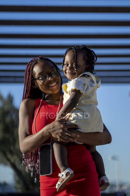 D'en bas de joyeuse mère afro-américaine avec des tresses et des vêtements élégants debout avec une petite fille souriante sur les mains dans la rue de la ville au soleil — Photo de stock