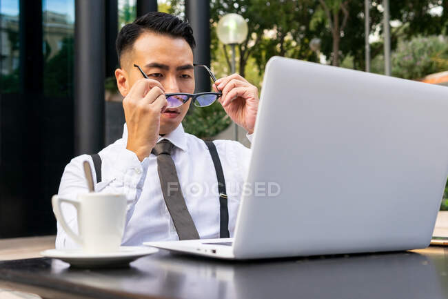 Empresario masculino étnico joven bien vestido poniéndose anteojos contra la mesa con netbook y bebida caliente en la cafetería de la calle - foto de stock