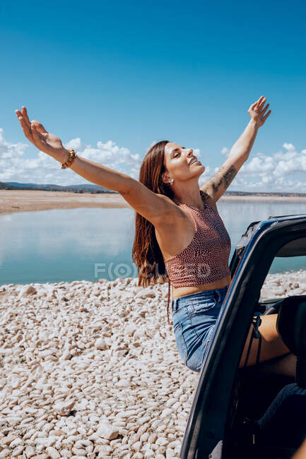 Side view of young female in denim jeans and top leaning out of car window with outstretch arms and eyes closed on coast of pond — Stock Photo
