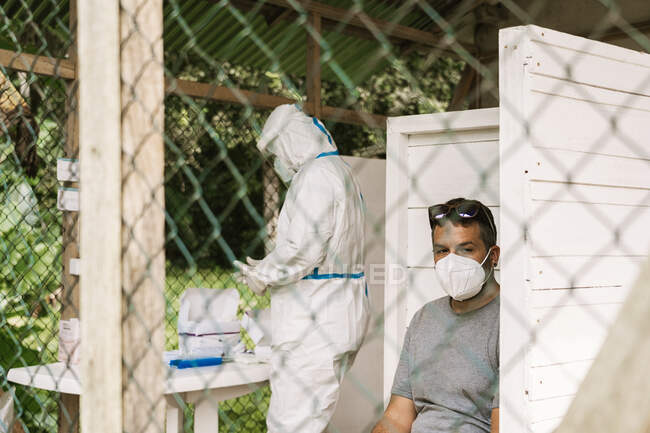 Through metal fence of side view of unrecognizable doctor in protective uniform standing and preparing vaccine injection for ethnic male in sterile mask sitting and looking away during COVID pandemic — Stock Photo