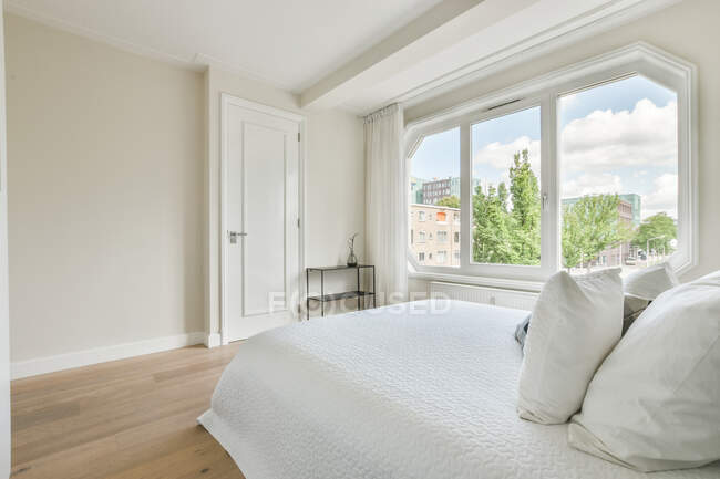 Cómoda cama con manta y almohadas situada cerca de la ventana en el dormitorio soleado de apartamento moderno - foto de stock