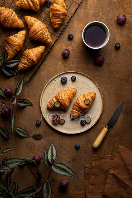 From above of tasty fresh baked croissants served on plate with fruits placed near cup of tea on wooden table in morning time in light room — Stock Photo