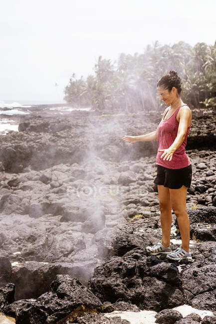 Vue latérale de la jeune voyageuse heureuse en vêtements décontractés debout près du geyser fumant avec les yeux fermés lors d'un voyage de randonnée à Sao Tomé — Photo de stock