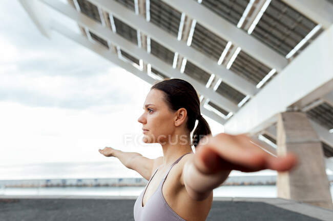 Mujer decidida en ropa deportiva con brazos extendidos haciendo ejercicio de yoga en la calle cerca del panel fotovoltaico moderno durante el entrenamiento en la ciudad - foto de stock